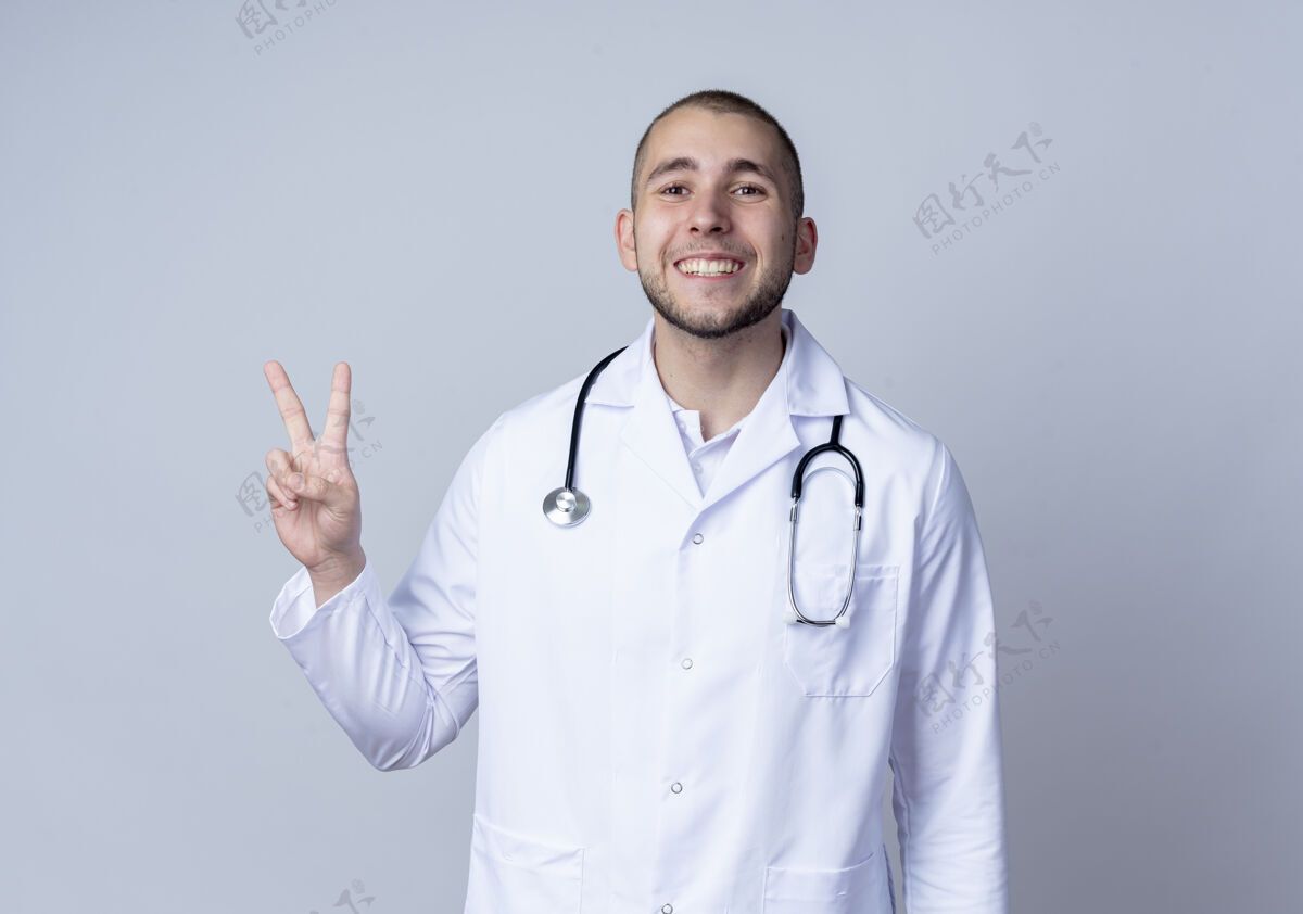 人面带微笑的年轻男医生身穿医用长袍 脖子上戴着听诊器 在白墙上做着与世隔绝的和平手势听诊器男性微笑