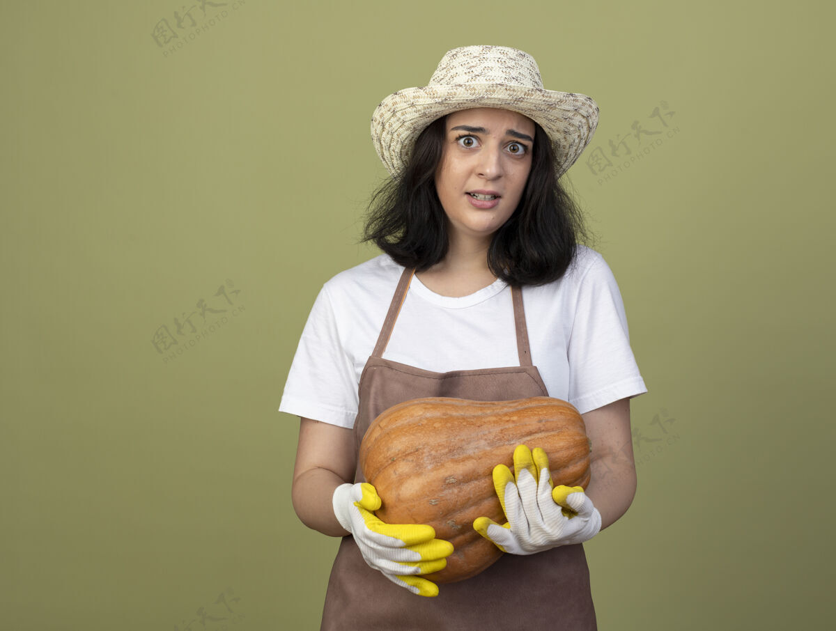 手套焦急的年轻黑发女园丁穿着制服 戴着园艺帽和手套 把南瓜孤立地放在橄榄绿的墙上穿脸橄榄