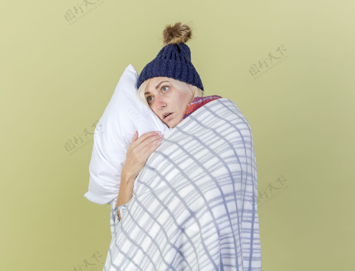 表情焦急的年轻金发病女人戴着冬天的帽子和围巾裹着格子呢抱枕看着隔离在橄榄绿墙上的一面帽子斯拉夫穿着