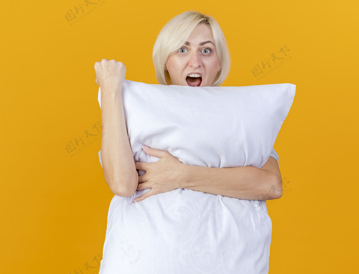 表情兴奋的年轻金发病女人抱住枕头 把拳头举起孤立在橙色的墙上市民人脸