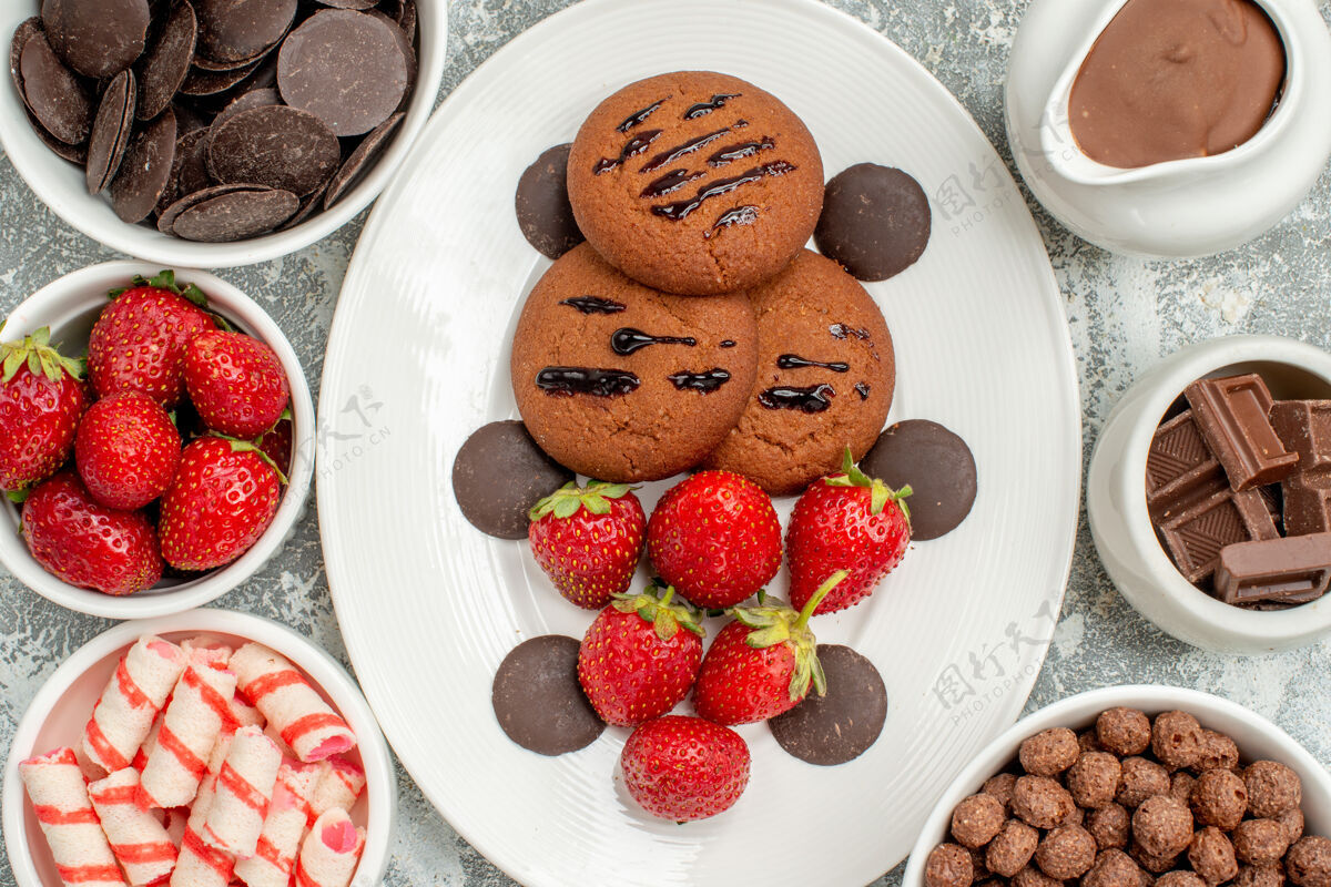 浆果顶部近距离观看白色椭圆形盘子上的巧克力饼干草莓和圆形巧克力 桌子上的碗里放着糖果草莓巧克力麦片和可可巧克力早餐盘子