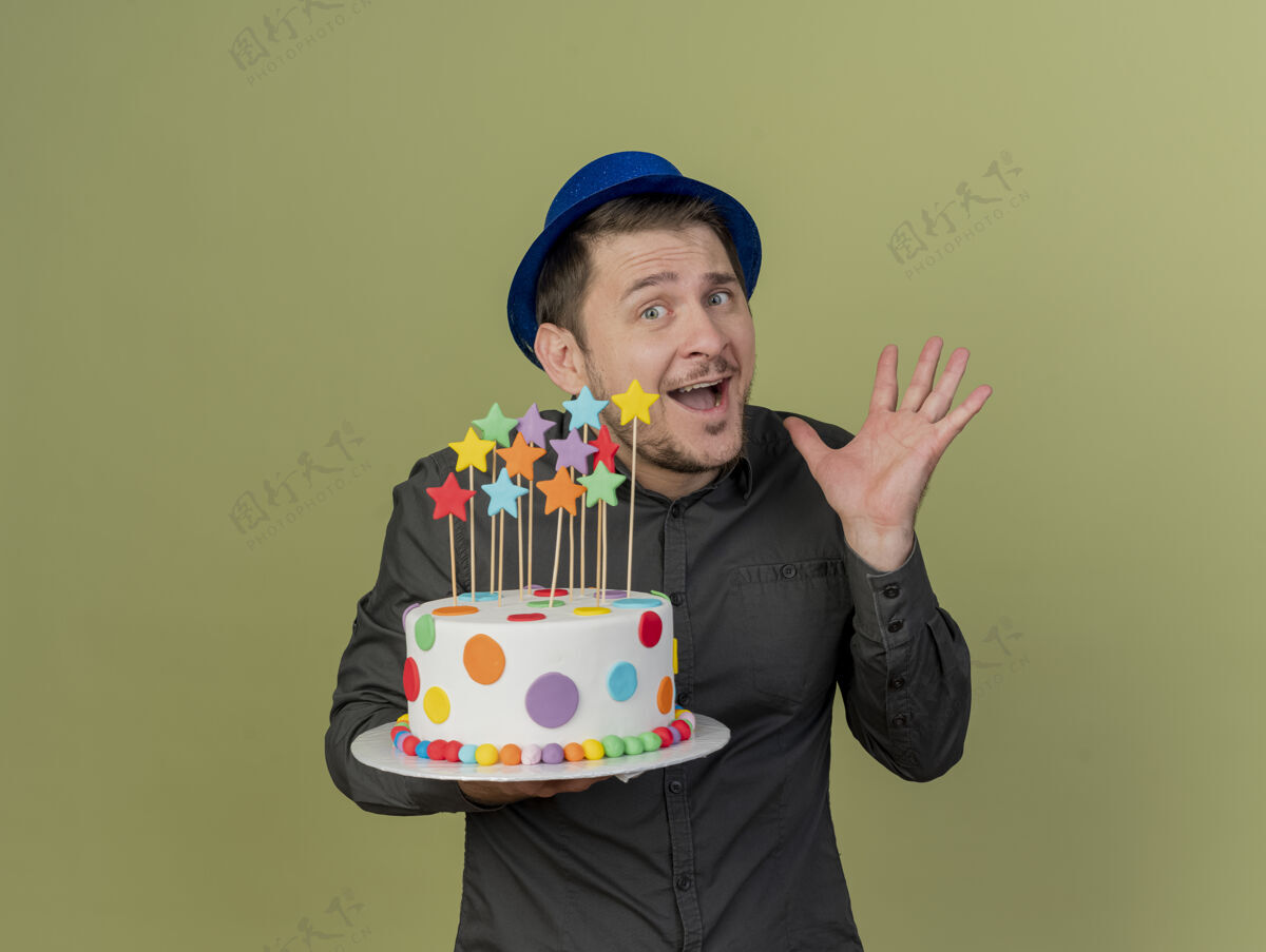 高兴快乐的年轻人穿着黑色衬衫 戴着蓝色帽子 手里拿着蛋糕 手被隔离在橄榄绿上派对黑色摊开