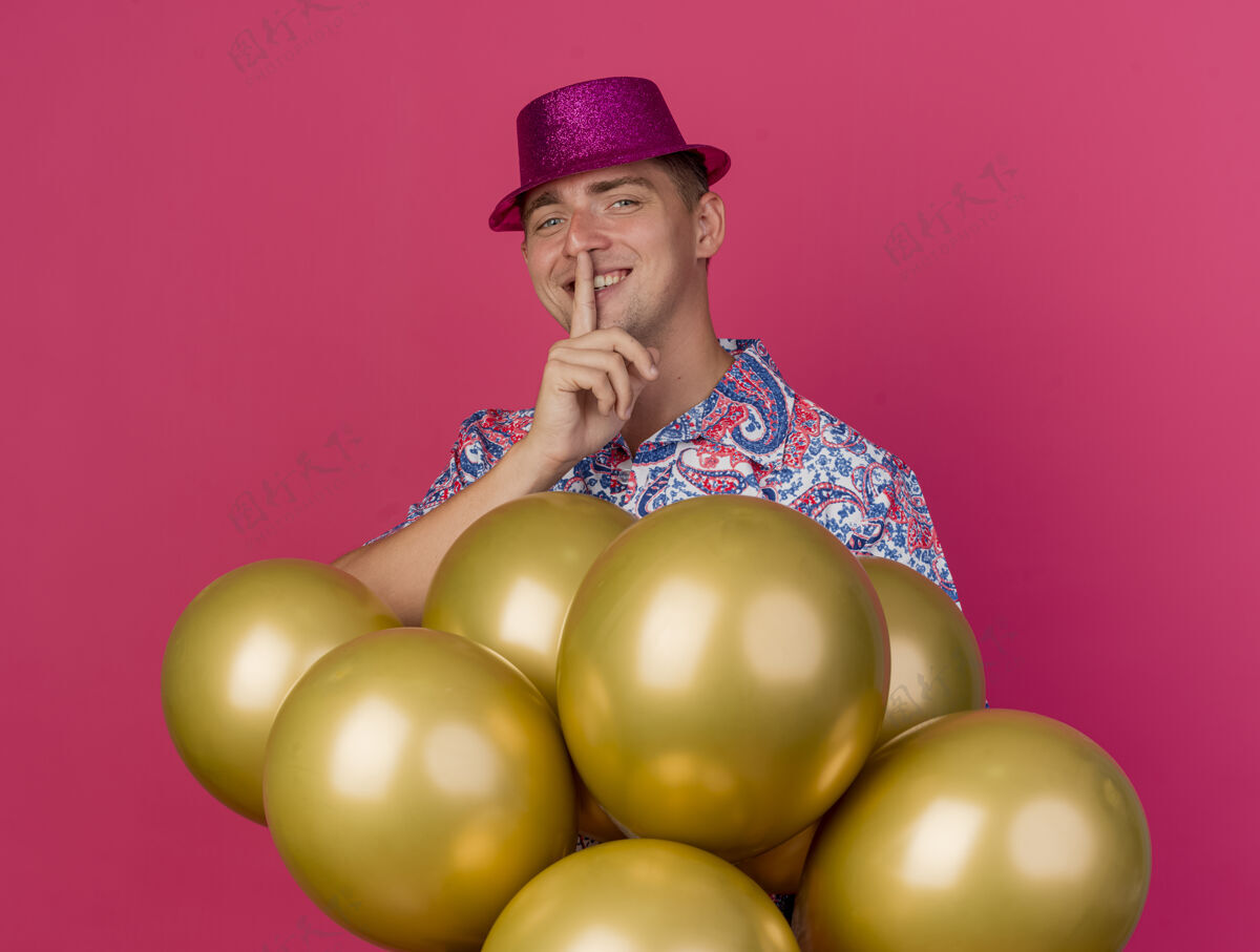 男人微笑的年轻人戴着粉红色的帽子站在气球后面 在粉红色的气球上表现出沉默的姿态站立手势派对