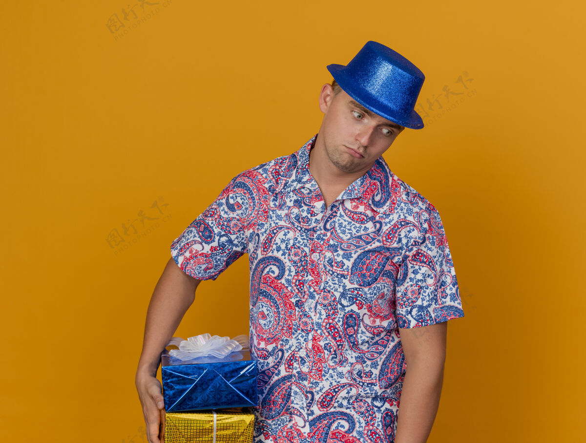 蓝色悲伤的年轻人戴着蓝帽子拿着礼品盒孤立在橙色的聚会年轻戴着帽子