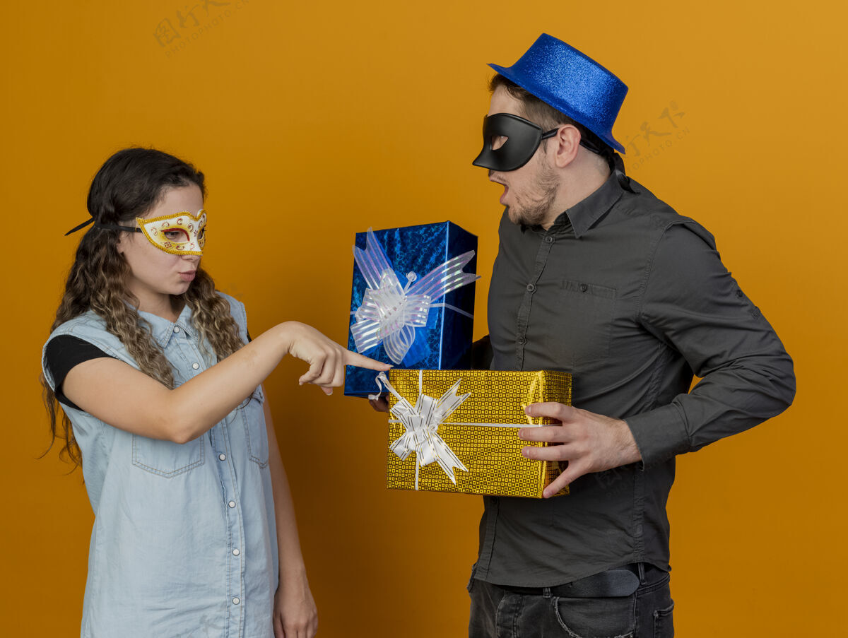 橙色年轻的派对女郎戴着化装眼罩把手指放在礼品盒上的男生手上孤立在橙色上派对化装舞会年轻人
