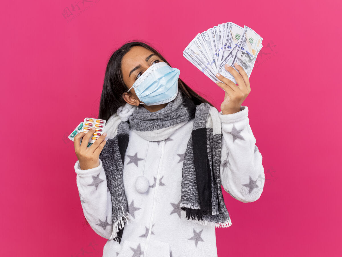 粉红色给人印象深刻的年轻病女孩戴着医用面罩 戴着围巾 拿着药片 看着手里的现金孤立在粉色上医疗印象围巾