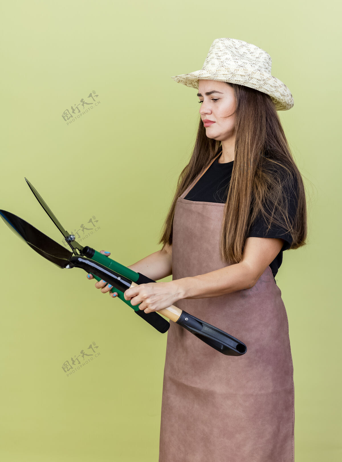 花园体贴美丽的园丁女孩穿着制服 戴着园艺帽 拿着剪刀看着橄榄绿上的铁锹园艺黑桃帽子