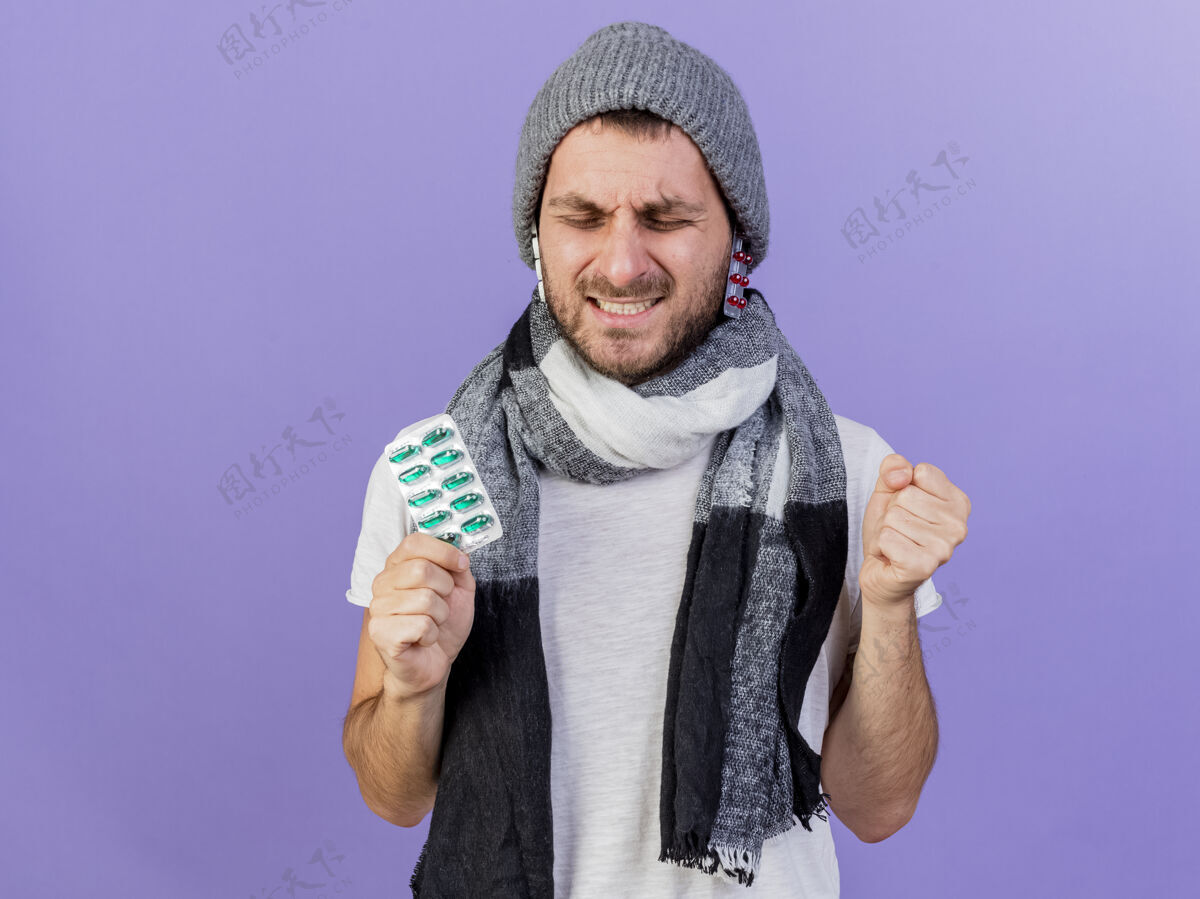 戴着快乐的年轻病夫戴着冬天的帽子 戴着围巾 帽子下面放着药丸 拿着药丸 在紫色上显示出“是”的手势药片男人快乐