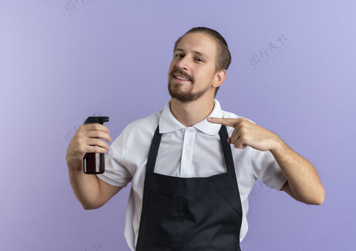 制服面带微笑的年轻英俊的理发师穿着制服 拿着并指着隔离在紫色墙上的喷雾瓶理发师微笑衣服