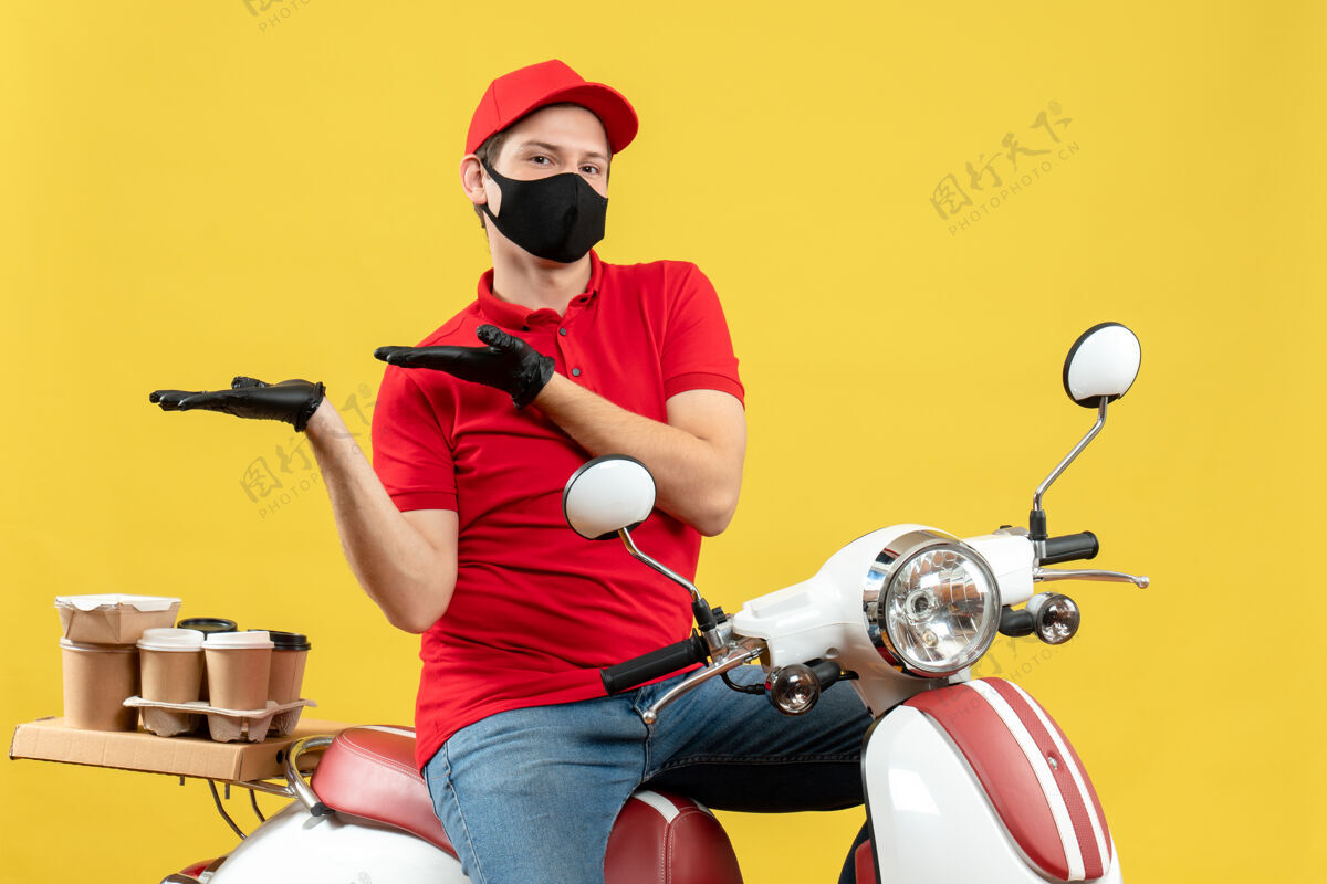 背景戴着医用面罩 穿着红色上衣 戴着帽子手套 坐在滑板车上 指着黄色背景上右边的东西 思考的年轻人的正视图车辆黄色右