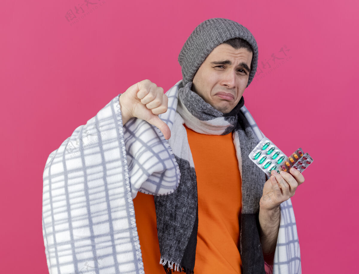 男人可怜的年轻人戴着冬天的帽子 围巾裹着方格布 手里拿着药片 大拇指朝下 孤零零地躺在粉红色的地板上疾病拇指放下