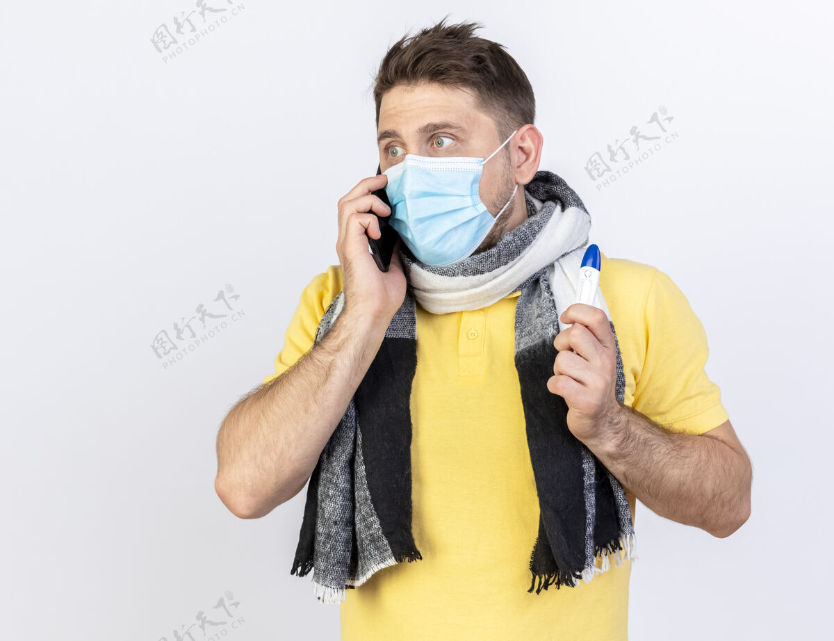 人焦急的年轻金发病患戴着医用口罩和围巾讲电话 拿着隔离在白墙上的温度计人人抱