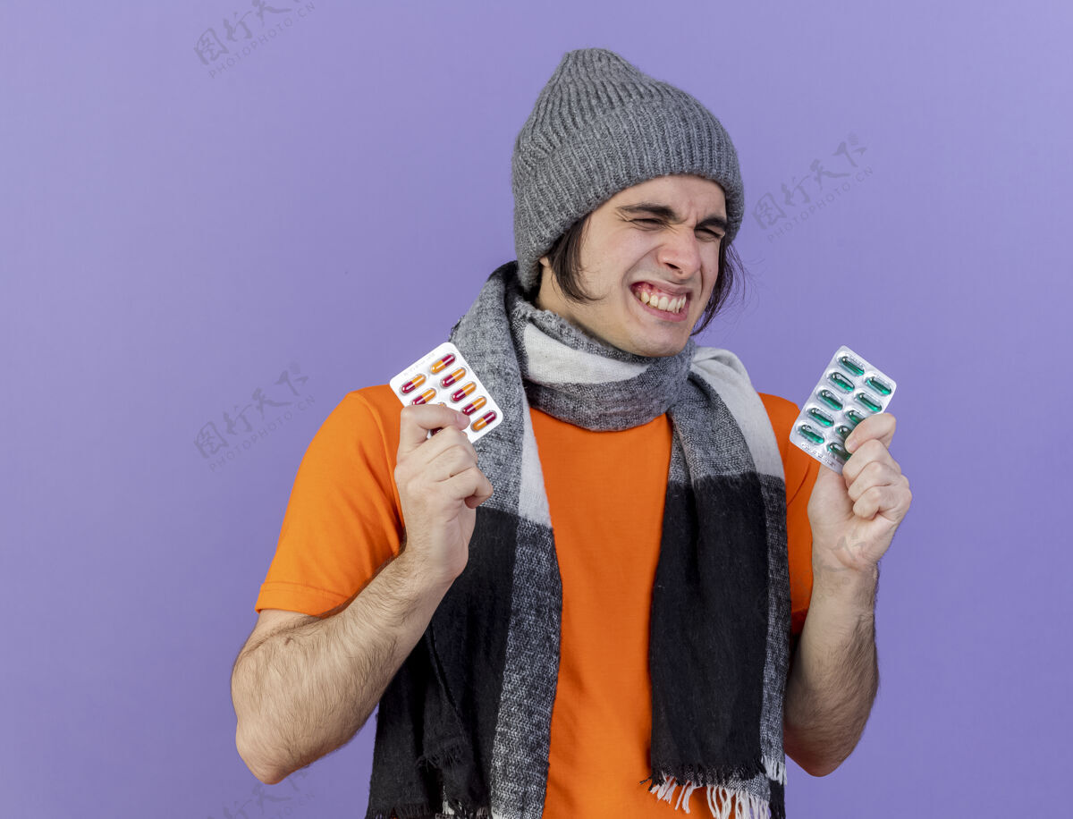 男人快乐的年轻病夫戴着冬天的帽子 戴着围巾 手里拿着紫色隔离的药丸围巾疾病眼睛