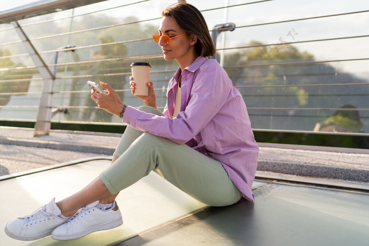 快乐时尚短发女子坐在摩登桥上 一边喝咖啡一边用手机城市休闲健康
