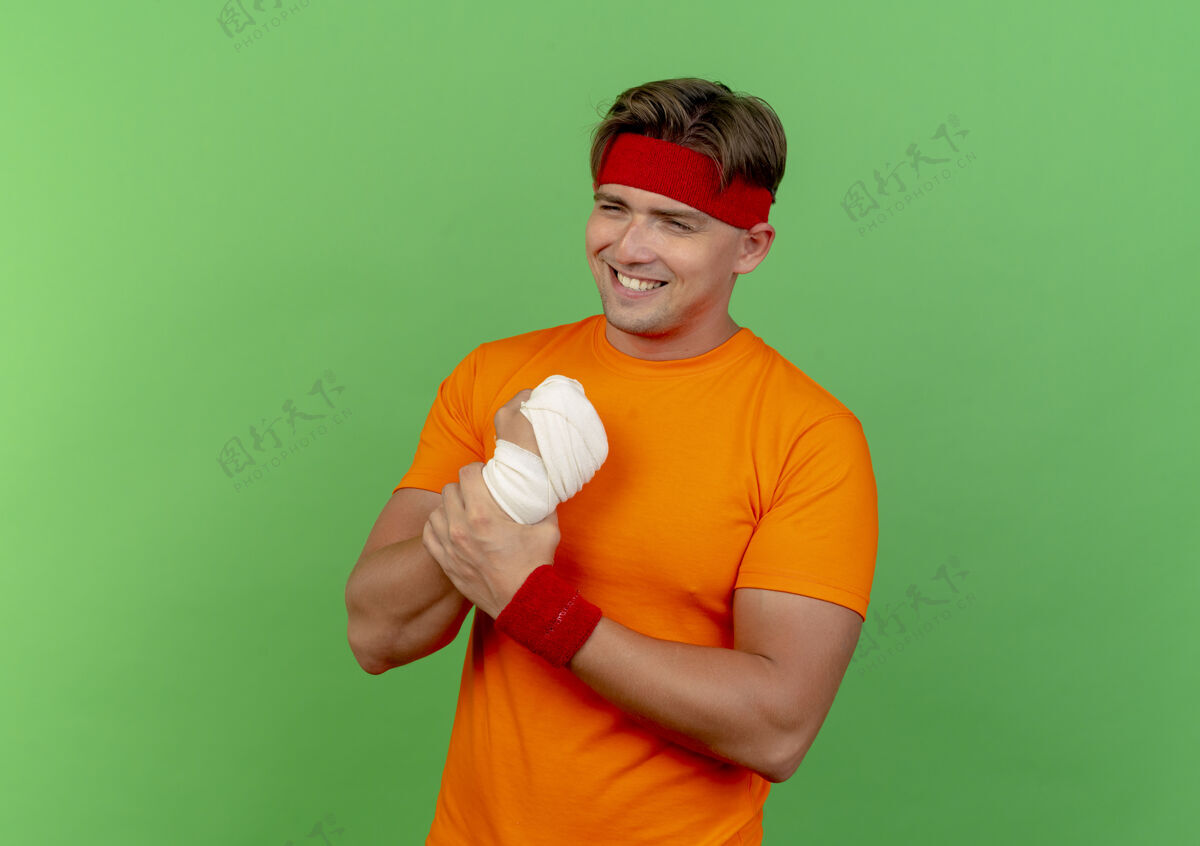 抱着面带微笑的年轻英俊的运动型男子戴着头带和腕带 用绷带包裹着他受伤的手腕 隔离在绿色的墙上市民绿色腕带