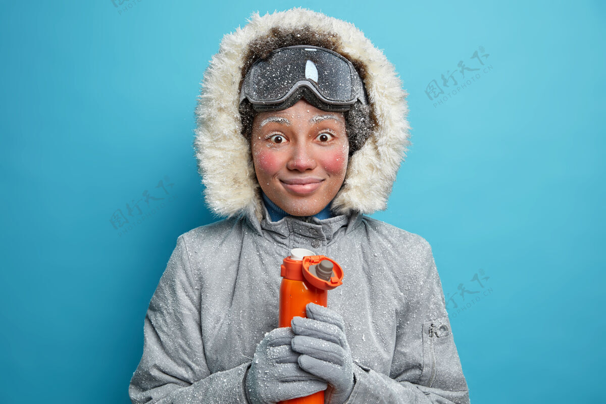 蓝色霜冻时面红耳赤的开朗冻女人微笑愉快地在滑雪活动后喝咖啡休息穿着暖和的冬衣拿着热饮料的保温瓶天气人满意