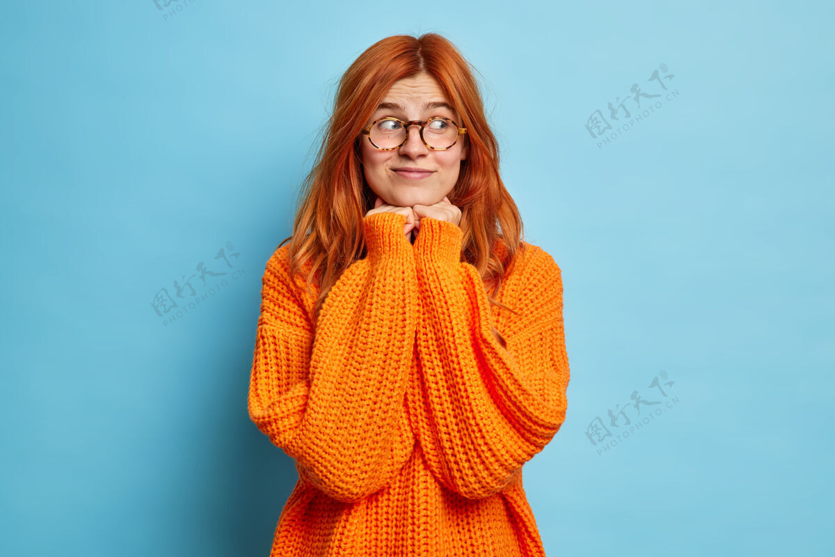 编织长着一头天然红发的年轻漂亮女人 手放在下巴下面 一边看一边想着一些愉快的事情 戴着眼镜 穿着橙色针织套头衫微笑高兴快乐