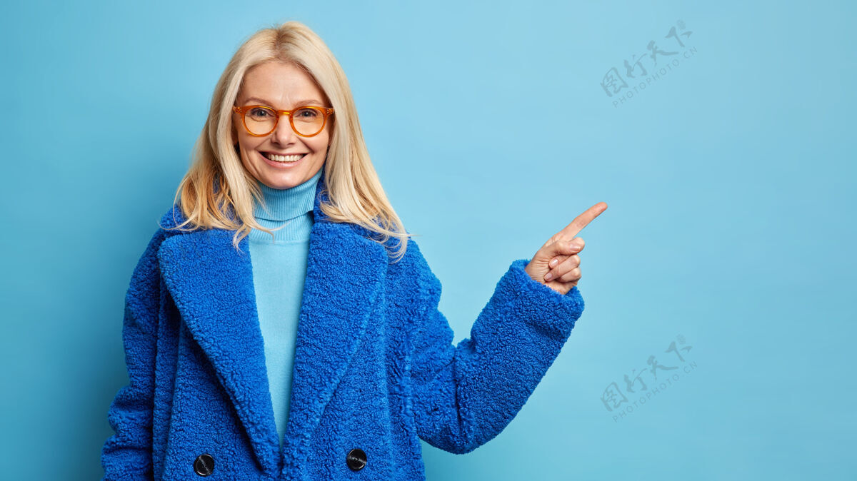 乐观美丽的金发中年妇女戴着眼镜 穿着温暖的蓝色外套 指着复印空间信任姿势外套