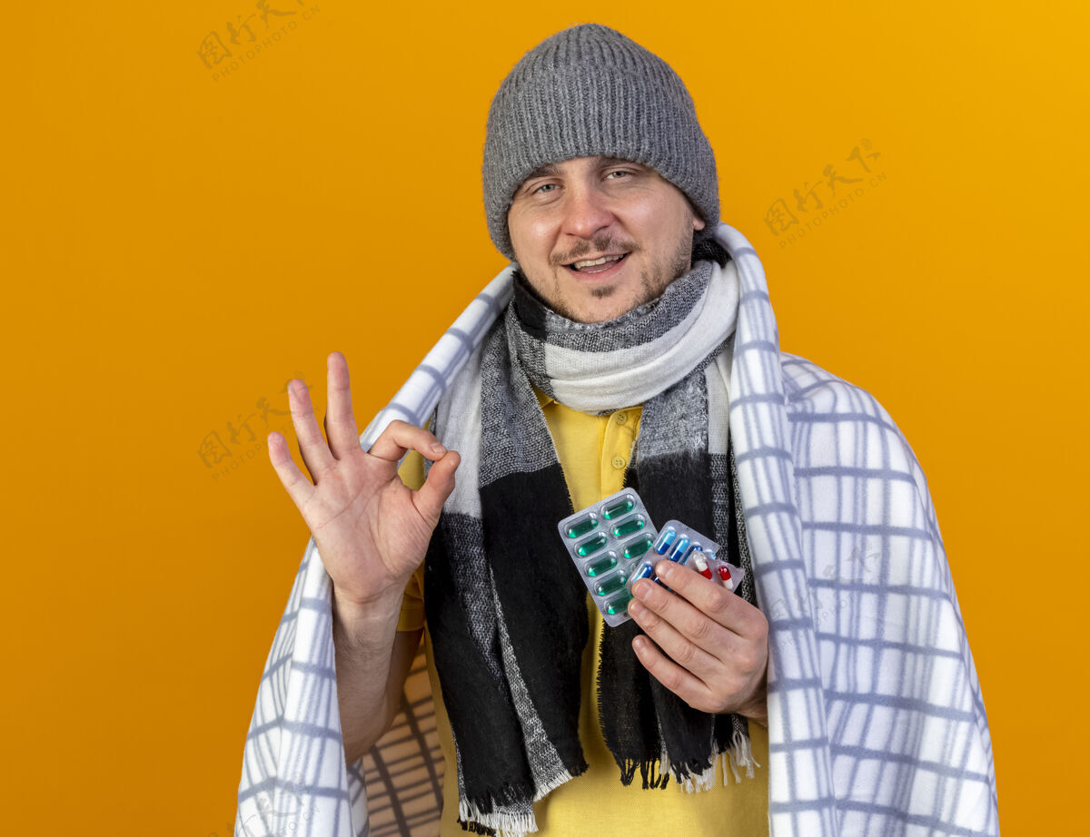 男人高兴的年轻金发病斯拉夫男子戴着冬天的帽子和围巾手势好的手势 并持有一包医药药片包裹在格纹隔离在橙色墙上的复制空间请包包裹