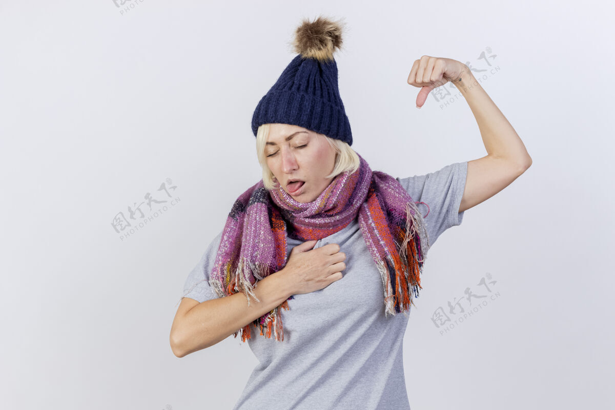 穿恼怒的年轻金发病斯拉夫妇女戴冬季帽子和围巾把拳头放在胸部和紧张的二头肌隔离在白色的墙壁与复制空间二头肌年轻胸部