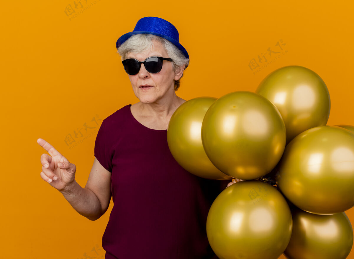 壁板戴着派对帽 戴着太阳眼镜的自信的老妇人站着 氦气球指向橙色墙壁上隔离的一面 还有复制空间帽子指点氦
