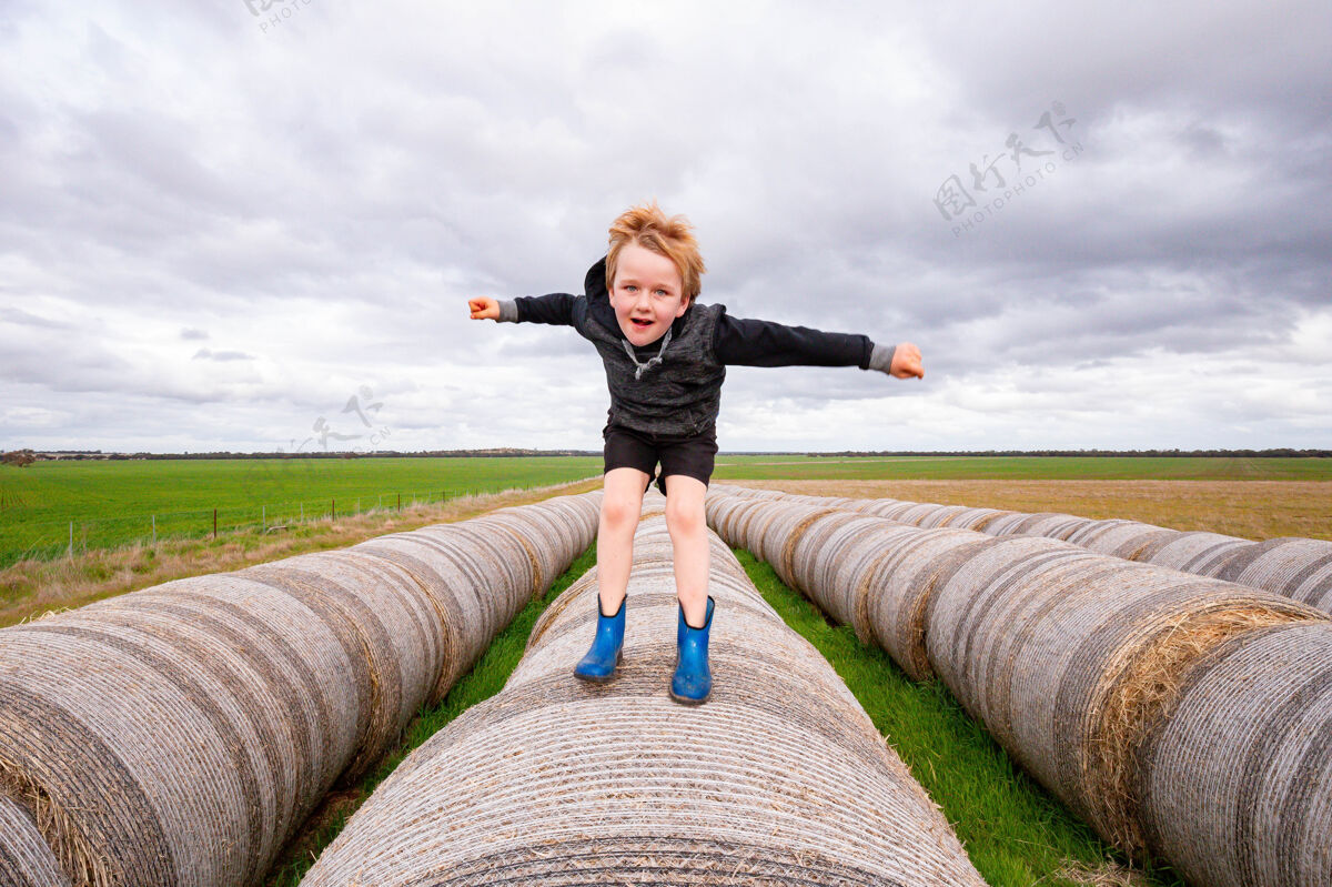 收获在阴天 金发碧眼的孩子在长长的一排圆形干草捆上跳跃——农场上的童年划船自然干草