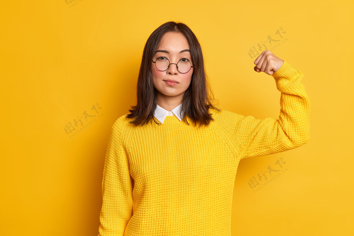 女性严肃的黑发亚洲女人的照片举起手臂 显示她的力量有强壮的肌肉站立自信的室内穿着黄色毛衣和圆眼镜女人的力量概念人呼吁二头肌