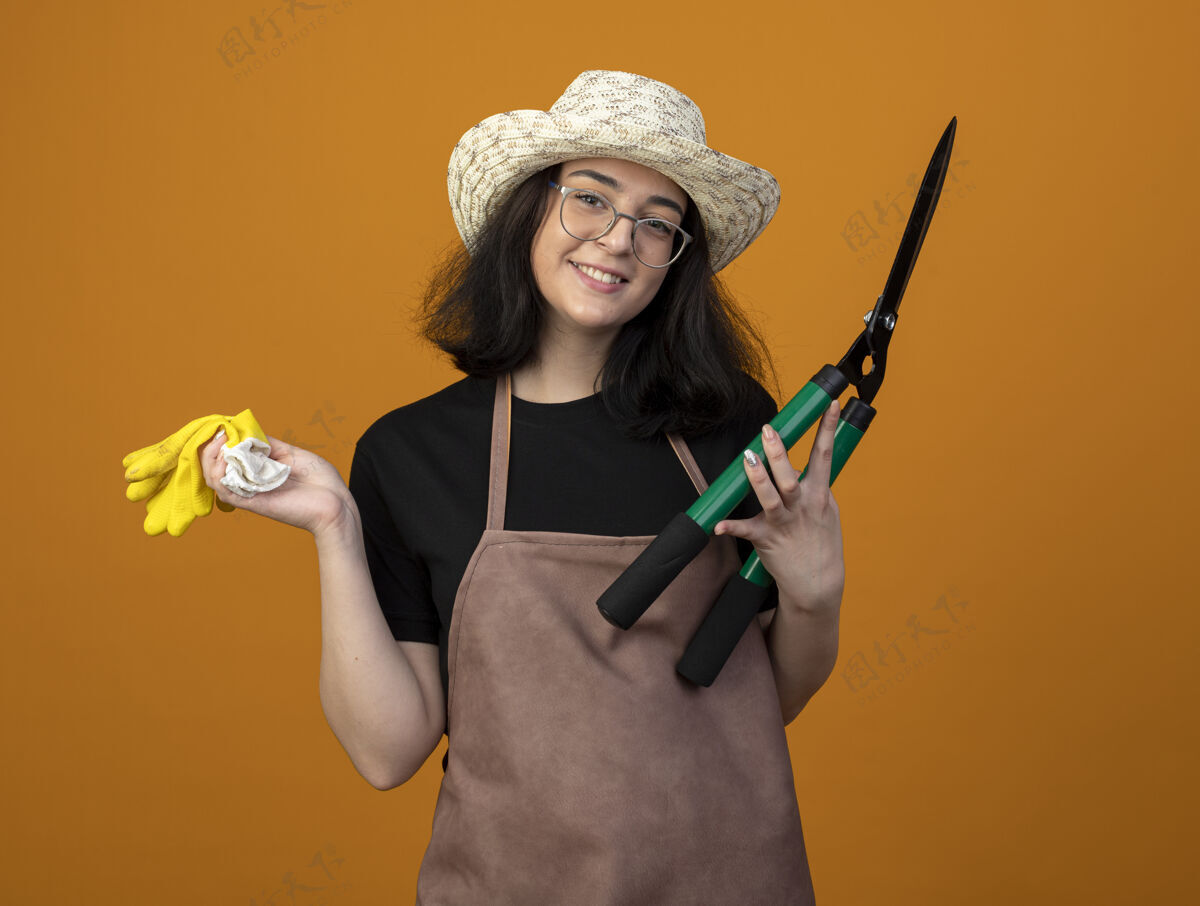 手套带着微笑的年轻黑发女园丁 戴着眼镜 穿着制服 戴着园艺帽 拿着园艺剪和手套 隔离在橙色的墙上 留着复印空间园艺剪子制服