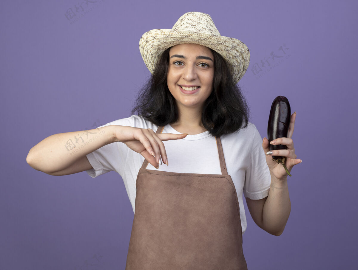 年轻人微笑着的年轻黑发女园丁穿着制服 戴着园艺帽 手持并指着隔离在紫色墙壁上的茄子 留着复制空间点园艺深色头发