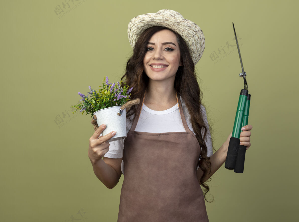 园艺微笑着的年轻女园丁穿着制服 戴着园艺帽 把花盆和花园剪子孤立地放在橄榄绿的墙上 留着复制空间橄榄举行穿