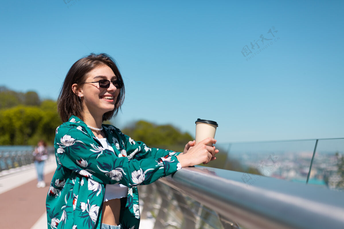 城市户外写真：穿着绿衬衫的女人 喝着咖啡 享受着阳光 站在桥上 清晨的城市景色令人惊叹观光茶城市景观