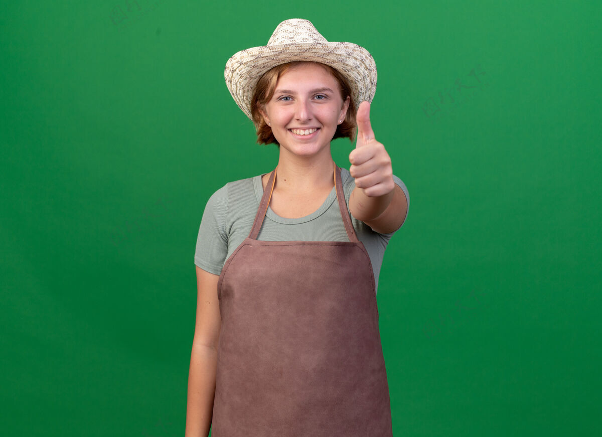 帽子微笑着的年轻的斯拉夫女园丁戴着园艺帽竖起大拇指在绿色斯拉夫人女的园艺