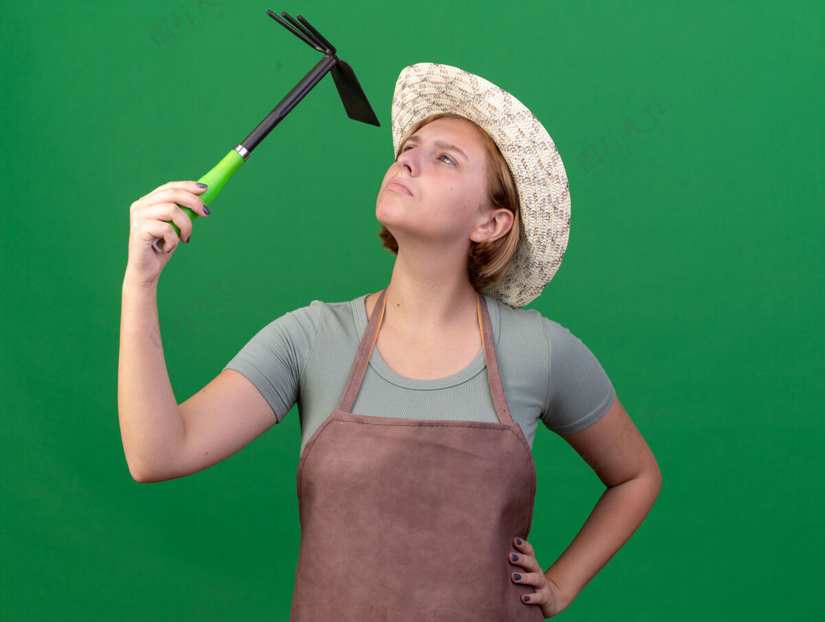 女的自信的年轻斯拉夫女园丁戴着园艺帽拿着锄头看着耙子上的绿色园艺斯拉夫人帽子