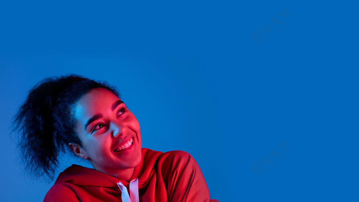 手耳机在五彩霓虹灯下的蓝色工作室背景上孤立的非洲裔美国女性肖像美丽的女性模特人类情感的概念 面部表情 销售 广告 时尚明亮面部脸