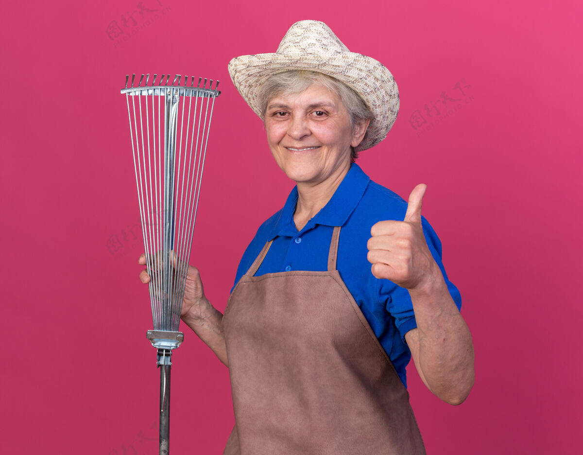 帽子微笑的上了年纪的女园丁戴着园艺帽 拿着耙叶 竖起大拇指对着粉色园艺雌性耙子