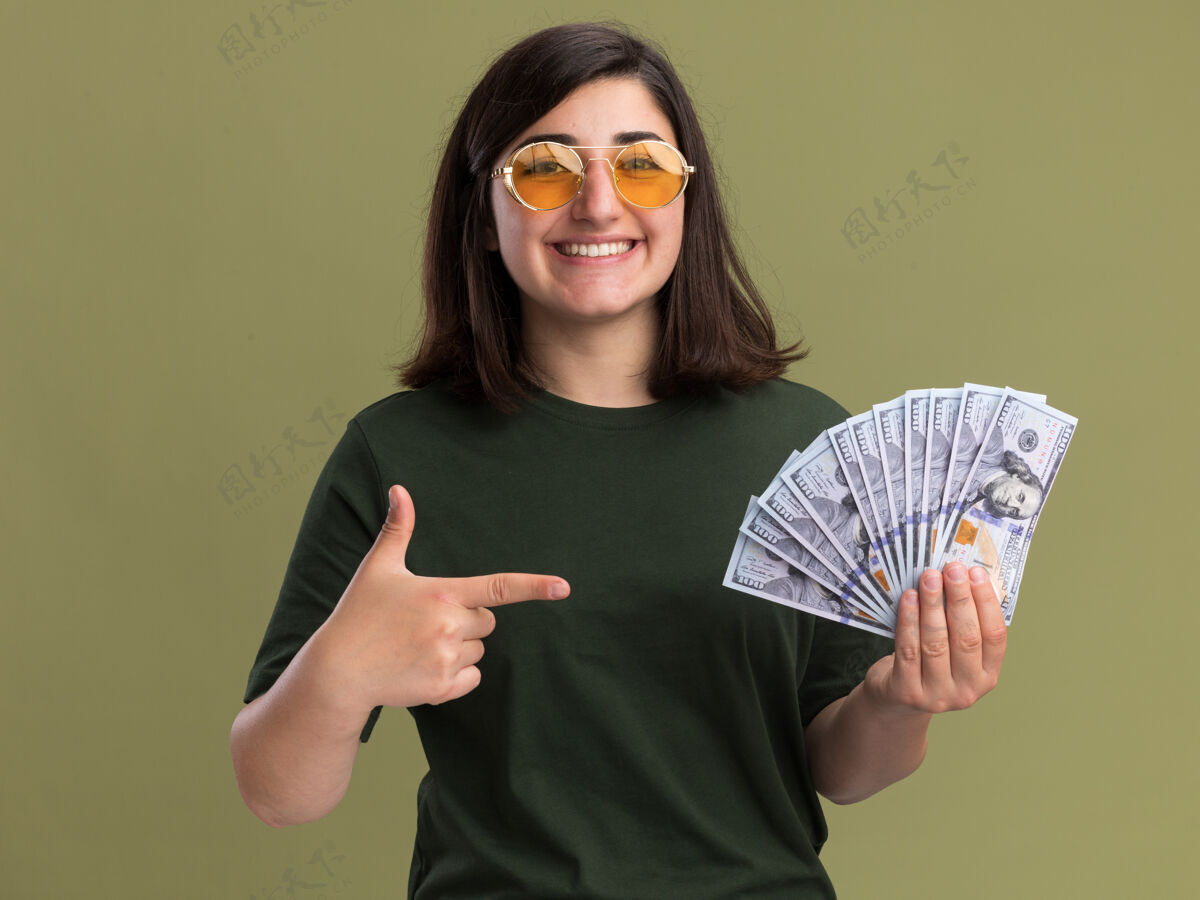指着微笑着的年轻漂亮的高加索女孩 戴着太阳眼镜 拿着钱 指着橄榄绿的相机漂亮抱着微笑