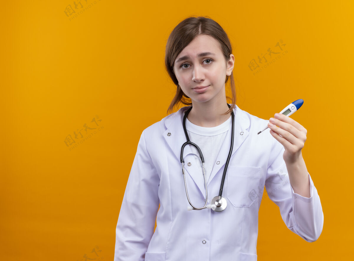 穿面容严肃的年轻女医生 穿着医用长袍 手持听诊器 拿着温度计 站在隔离的橙色空间里 还有复印空间长袍复制严重
