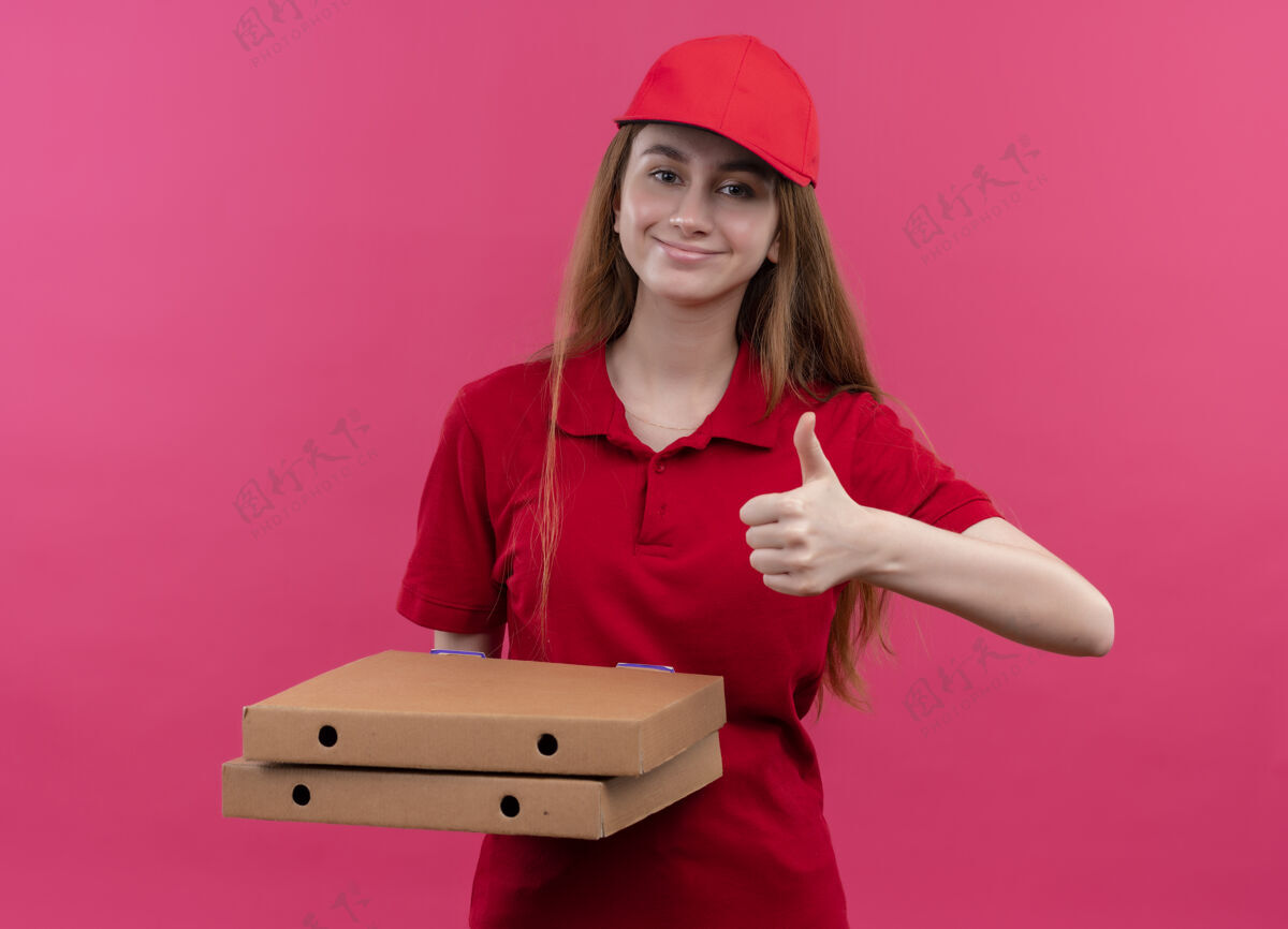 请穿着红色制服 拿着包裹 在孤立的粉色空间里竖起大拇指的年轻送货女孩很高兴盒子送货粉色
