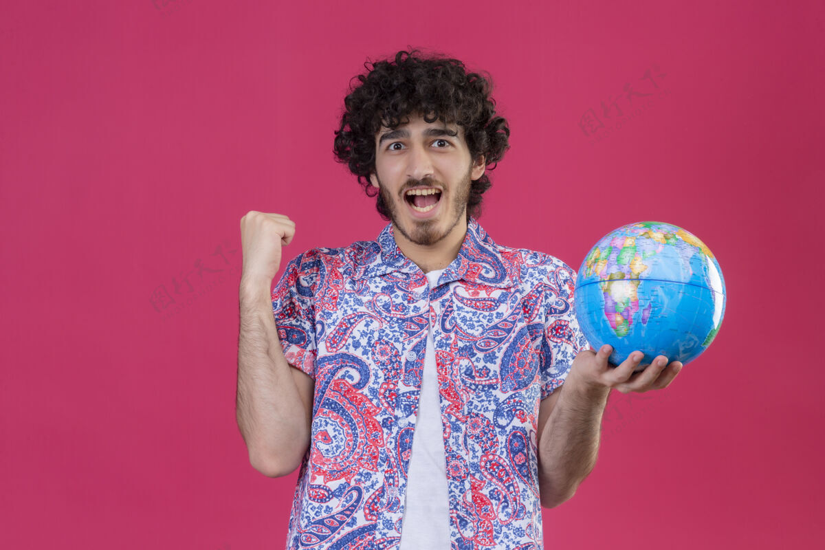 旅行者快乐的年轻英俊的卷发旅行者手持地球仪举起拳头在孤立的粉红色空间和复制空间提高快乐地球仪