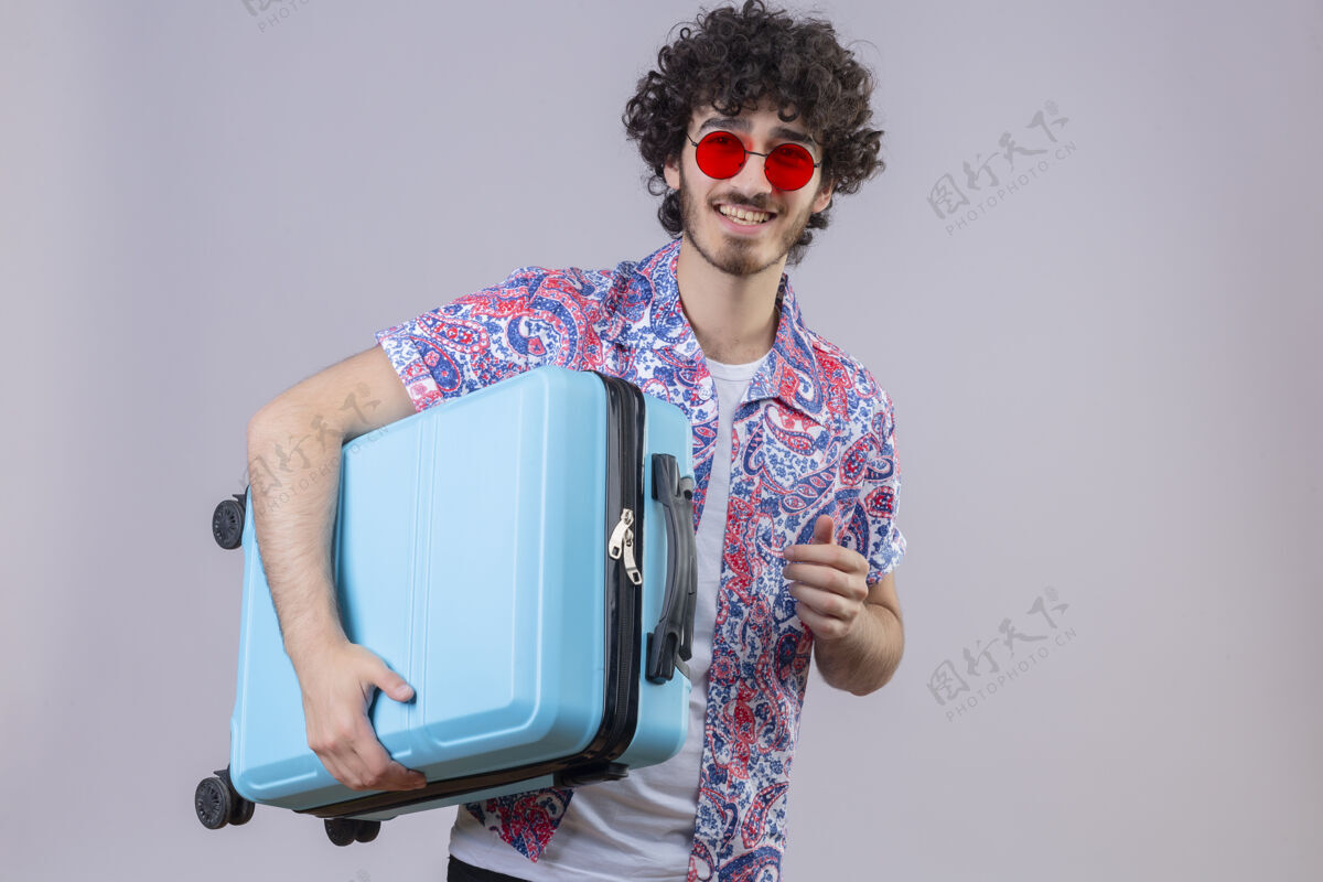 复制快乐的年轻帅气的卷发旅行者戴着墨镜拿着手提箱在与世隔绝的白色空间里复制空间太阳镜戴着旅行者