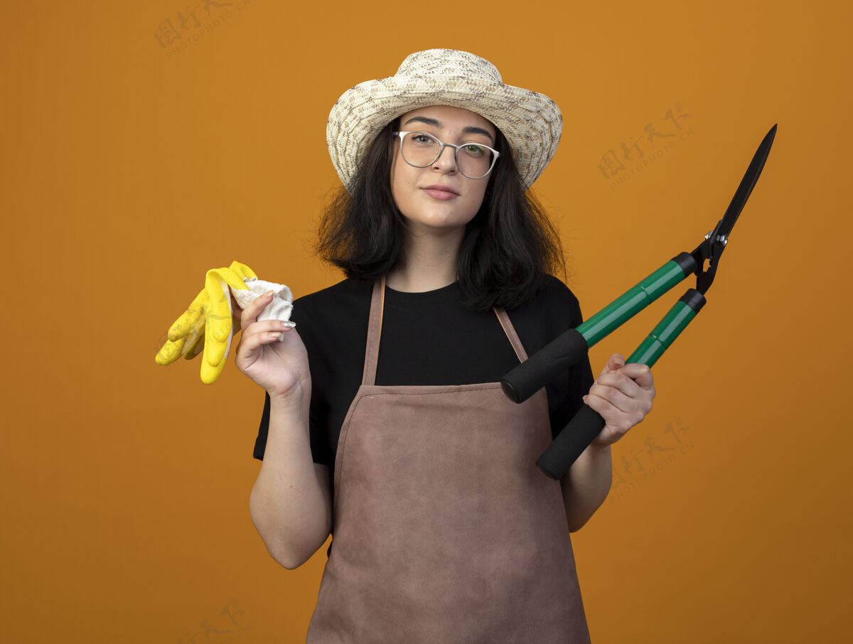 花园自信的年轻黑发女园丁 戴着眼镜 穿着制服 戴着园艺帽 拿着园艺剪和手套 隔离在橙色的墙上 留着复印空间年轻剪子光学