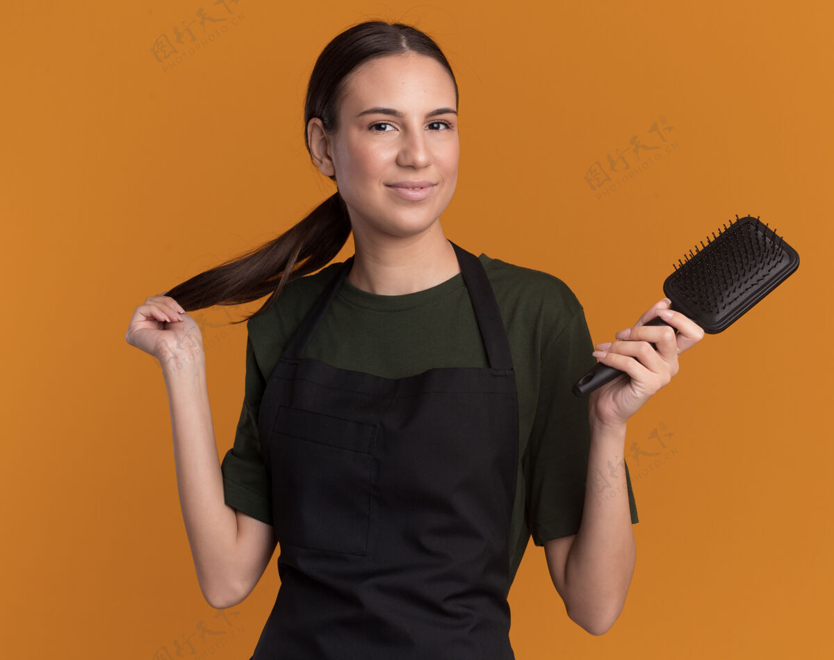 深色头发穿着制服的年轻黑发理发师女孩高兴地拿着发辫和梳子在橘子上梳子制服辫子