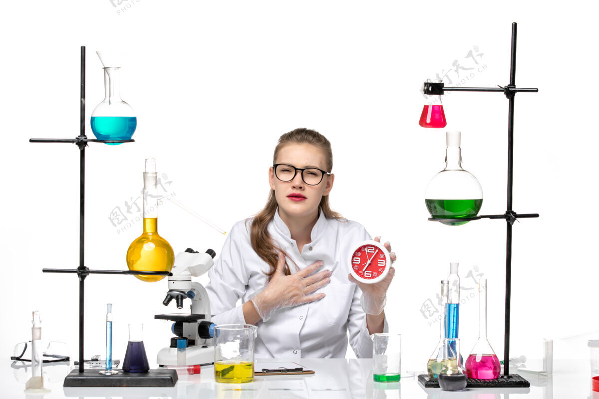 专业前视图穿着白色医疗服的女医生在白色背景上坐着拿着钟病毒化学大流行病毒科学钟诊所
