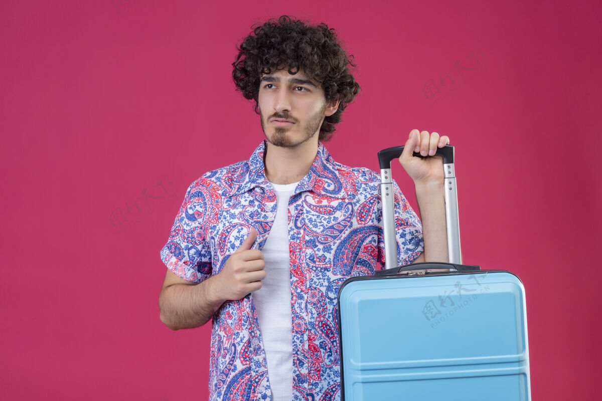 卷发自信的年轻帅气的卷发旅行家男人拿着手提箱 把手放在衬衫上 在一个孤立的粉色空间里 有一个复制空间男人自信手