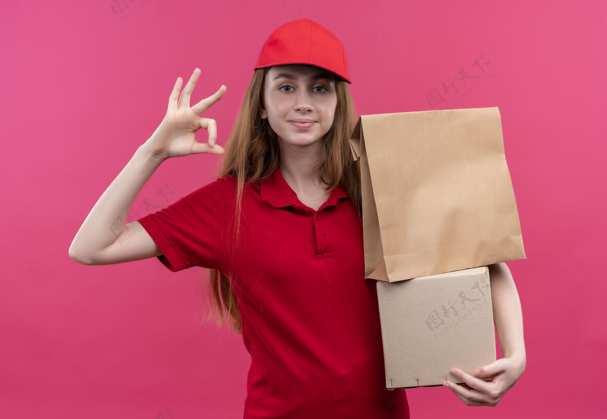 持有自信的年轻送货女孩穿着红色制服拿着箱子 在孤立的粉色空间上做着ok的标志信心制服好