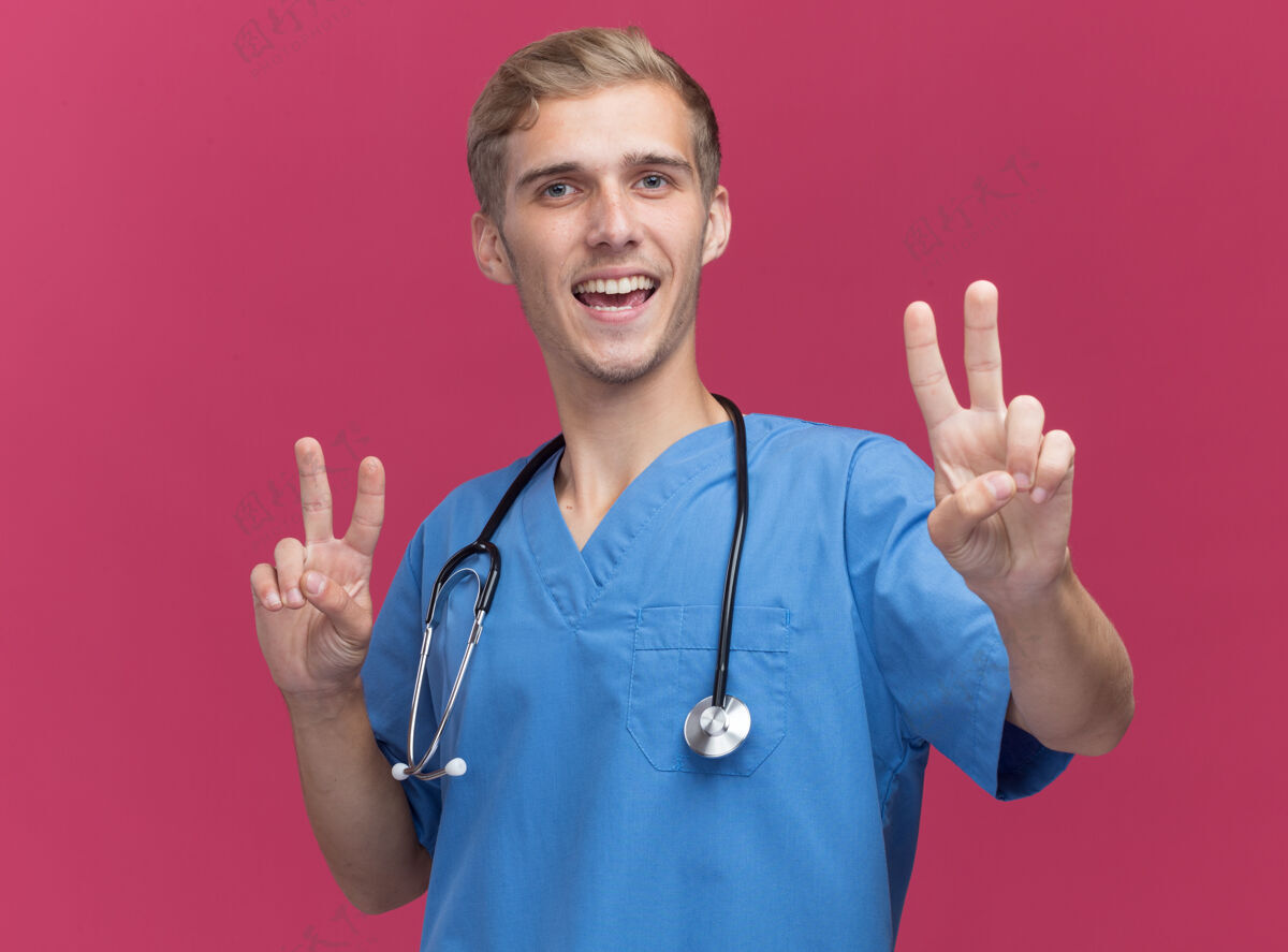 站立微笑的年轻男医生穿着医生制服 带听诊器 在粉红色的墙上显示出和平的姿态穿着表演表情
