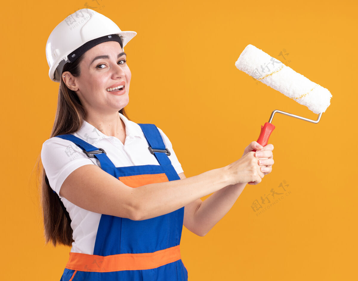 抱着身着制服的年轻建筑妇女微笑着拿着滚轴刷隔离在橙色的墙上微笑年轻人建设者