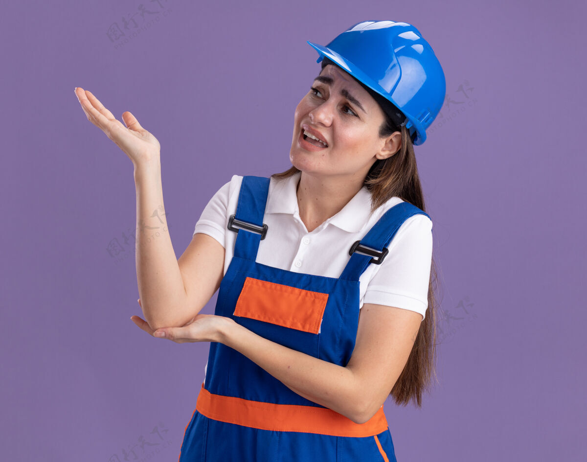 手不高兴地看着边上的年轻建筑工人妇女点着制服 手在边上孤立地站在紫色的墙上年轻立场姿势