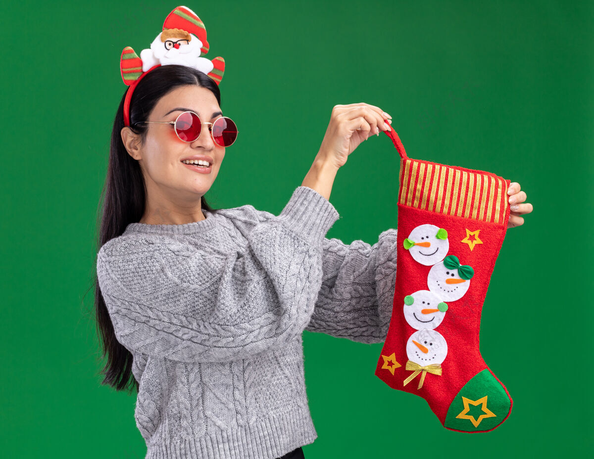 女孩快乐的年轻白人女孩戴着圣诞老人的头带 戴着眼镜 拿着圣诞长袜 看着隔离在绿色背景上的相机股票头带绿色