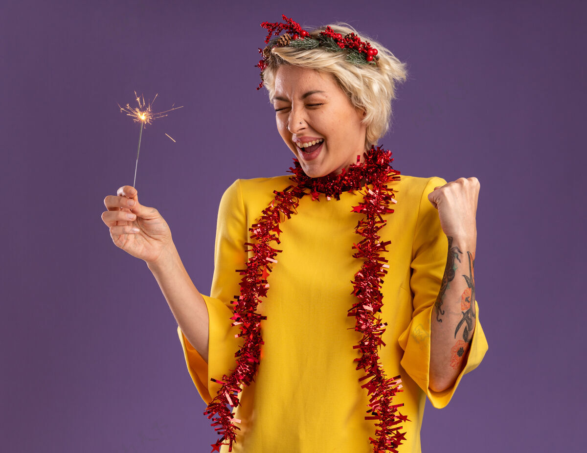 年轻快乐的金发女郎戴着圣诞花环 脖子上戴着金箔花环 手里拿着节日火花灯 做着“是”的手势 闭着眼睛 孤立地站在紫色背景上是女人关闭